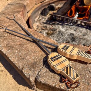 A close-up view of two Moose Tracks S'mores Sticks with a bonfire in the background. Chrome-plated s’mores sticks with light tan wooden handles shaped like ice cream cones. The handles are engraved with "Moose Tracks" in black lettering and finished with a leather loop at the base.